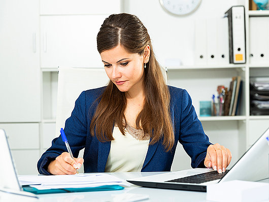 Eine Dame sitzt an einem Schreibtisch mit ihrem Laptop und ihren Dokumenten.
